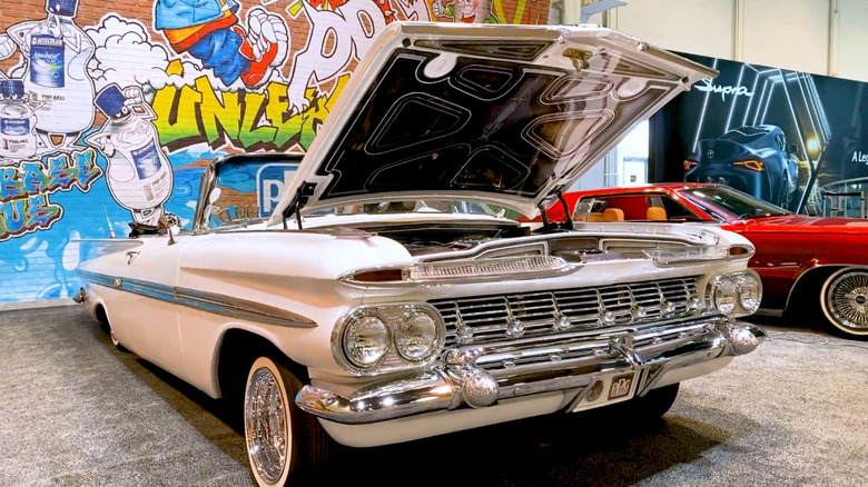 A classic white lowrider with its hood open on display at an indoor car show.
