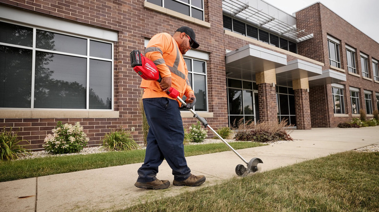 A man in an orange work shirt uses a Quik-Lok edger to clean up the grass along the sidewalk in front of a brick building.