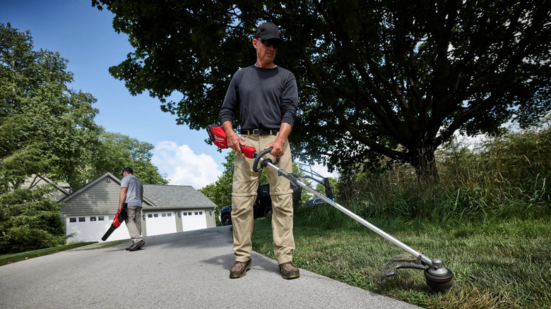 A man trimming grass with a Quik-Lok String Trimmer while another person uses a blower in the background.