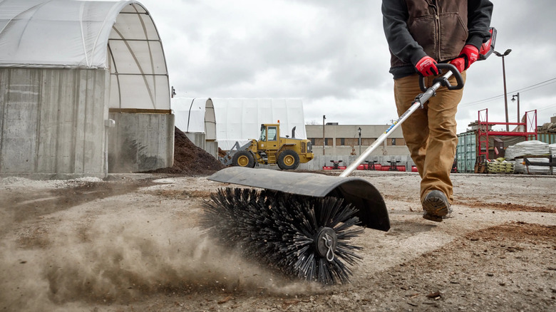 A person uses a Quik-Lok bristle brush to clear loose dirt off a hard path.