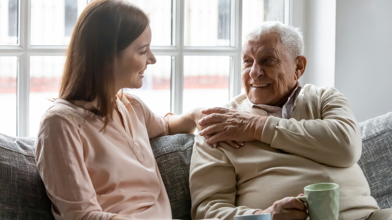 A smiling woman sitting on a couch with an older man with her hand on his shoulder