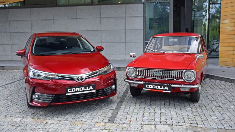 An old and a modern red Toyota Corolla parked in front of a modern-looking building with cobblestones.