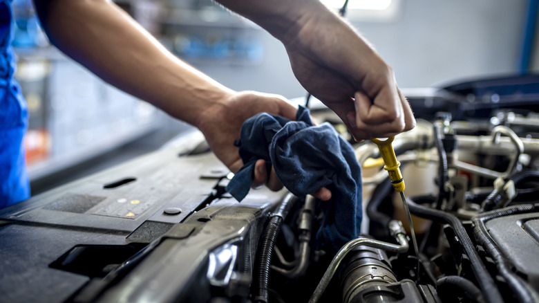 A mechanic holding a blue rag and an oil dipstick above a car with its hood open.