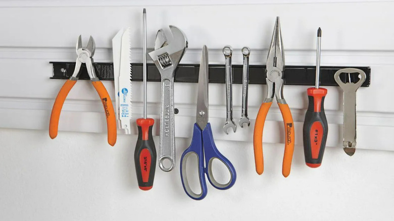 A magnetic tool bar from Harbor Freight screwed to a wall with pliers, razors, screwdrivers, spanners, scissor, wrenches, and a bottle opener on it.