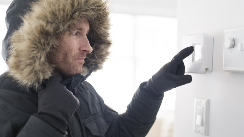 A man wearing a winter coat with a fuzzy hood and gloves adjusting an indoor thermostat