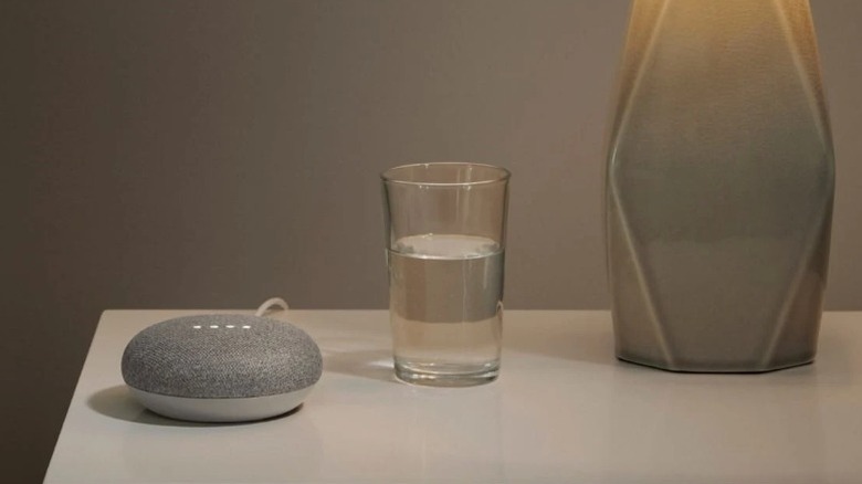 Google Nest Mini speaker on table next to glass of water and table lamp