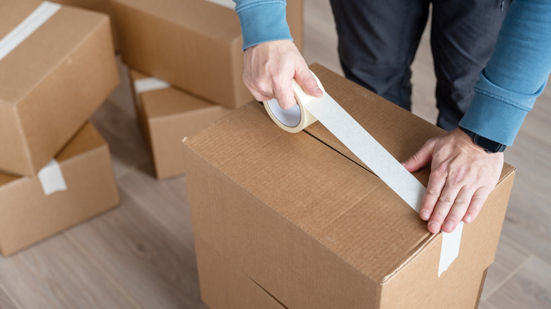 A man in a blue shirt using tape to close up a moving box, with more cardboard boxes in the background