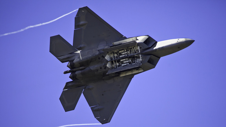 An F-22 Raptor in the air against a blue sky, with open missile docks