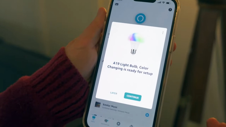 Hand holding phone with Alexa app showing Amazon Basics Smart Light Bulb ready for setup popup