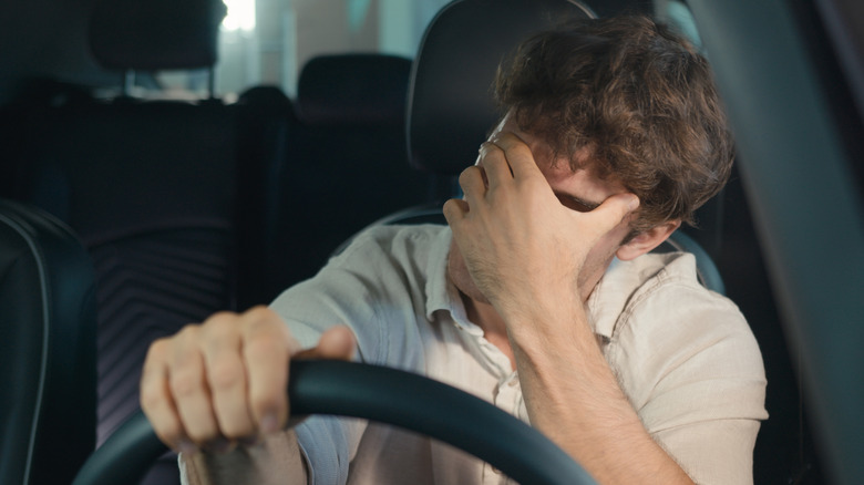 A frustrated person gripping the steering wheel with one hand, the other hand covering their face.
