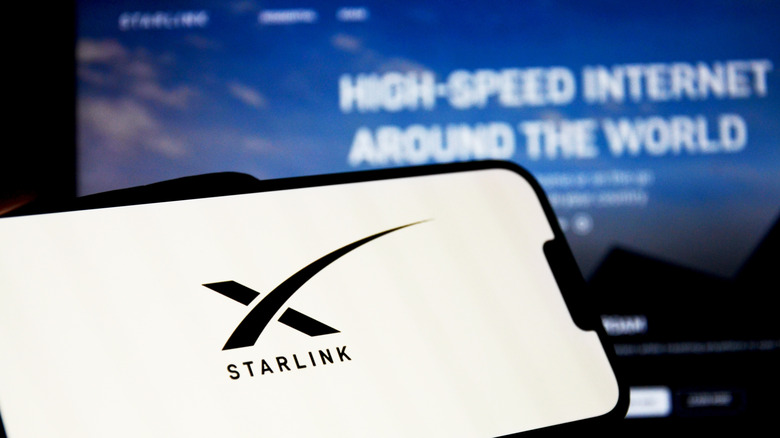 A phone with the Starlink logo in black on a white background sitting in front of a monitor with the Starlink website displayed
