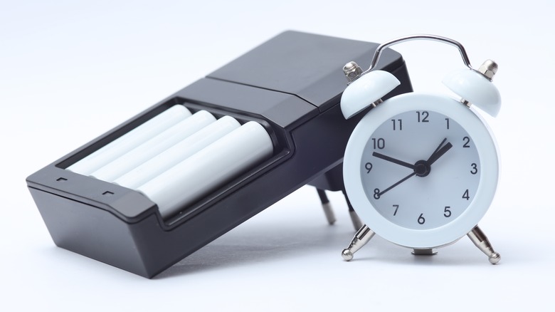 A generic black battery charger holding four white AA batteries next to a white analog alarm clock
