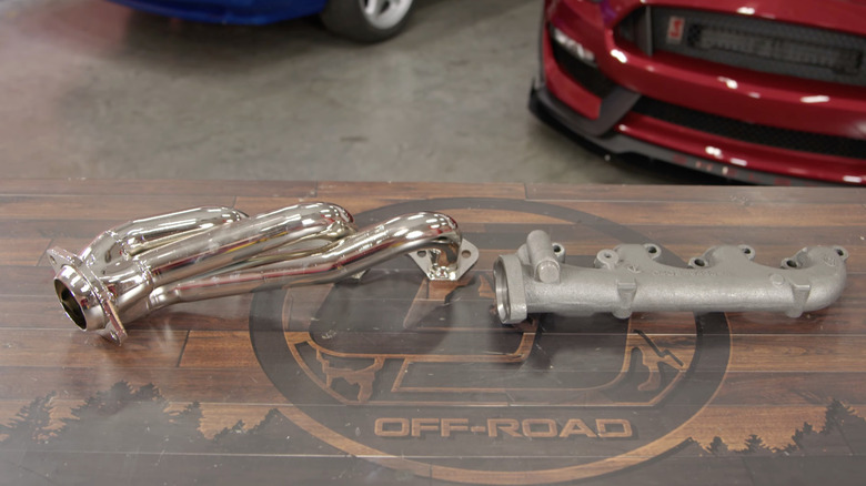 Polished short-tube headers next to a standard exhaust manifold on a wooden table, red Shelby GT350 in the background