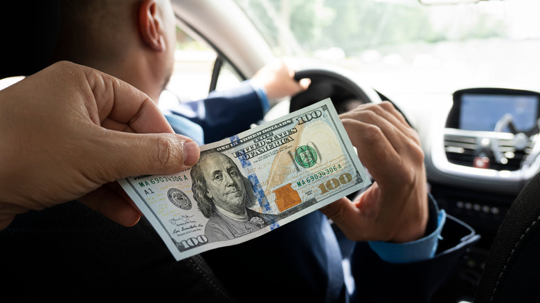 A person in the back seat of a car handing a $100 bill to the driver.