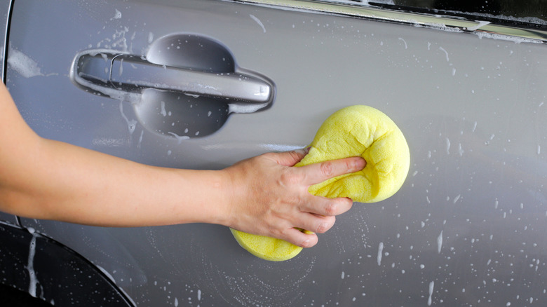 A hand using a yellow sponge to clean the side of a car with soap water.