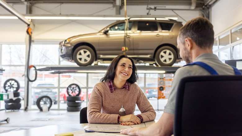 Female customer and male automotive service technician speaking at table inside automotive maintenance shop with SUV on lift in the background