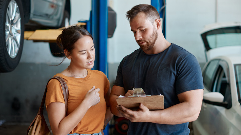Woman with automotive technician inspecting documents with cars on lifts in background