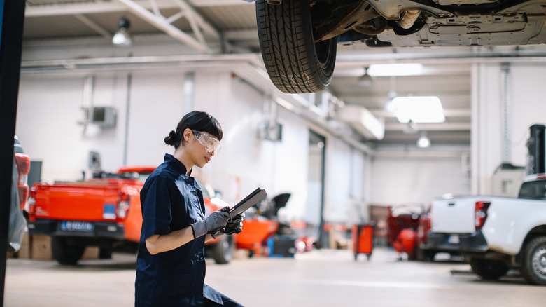 Female service technician at work in garage with pickup trucks in background and car's undetray on lift in foreground