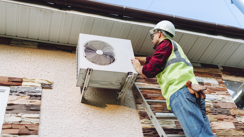 A technician in uniform standing on a ladder to repair an air conditioner.