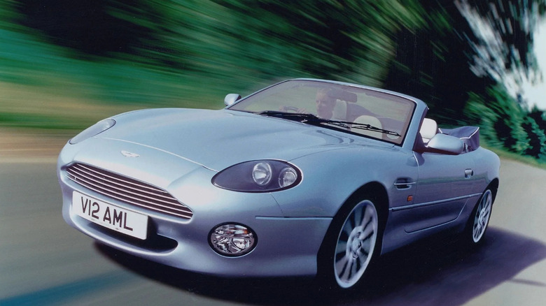 Silver Aston Martin DB7 Vantage Volante convertible driving at speed through a forested road, showcasing open-top luxury and British V12 performance.