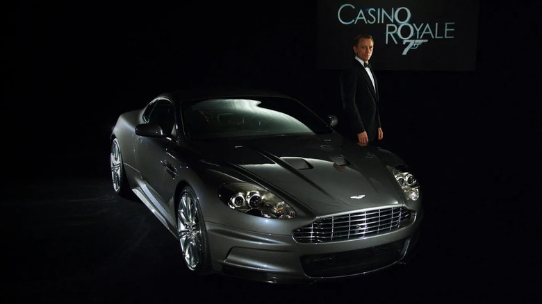 Aston Martin DBS from James Bond's Casino Royale showcased in a dark studio alongside a tuxedo-clad man, highlighting the car's cinematic elegance and performance.