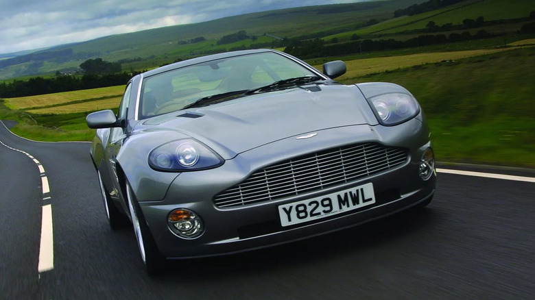 Silver Aston Martin Vanquish cruising on a scenic country road, highlighting its aggressive front fascia and grand touring performance under overcast skies.