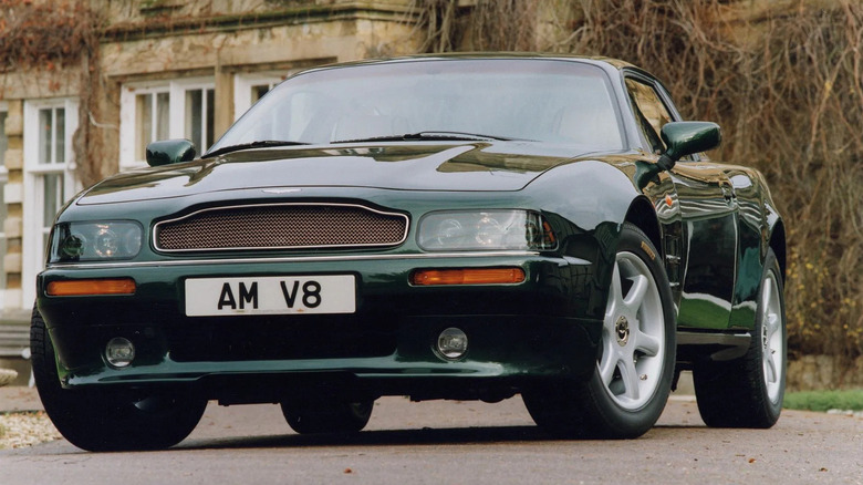 Dark green Aston Martin V8 Coupe parked in front of a historic stone mansion, highlighting classic British luxury and performance styling.