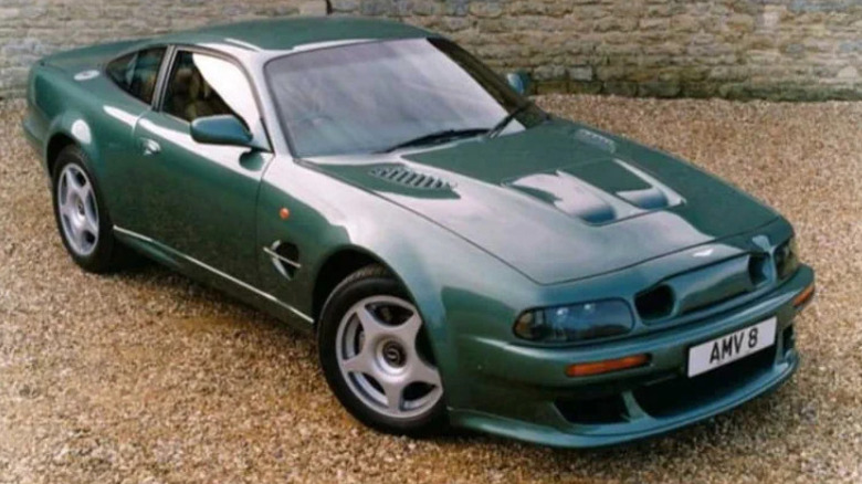 Aston Martin V8 Vantage in metallic green parked on gravel, featuring aggressive front air intakes and hood vents, showcasing 1990s British muscle styling.