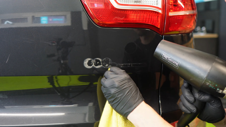 A person using a hairdryer and microfiber cloth to remove the EcoDynamics badge off an older black Kia Sportage