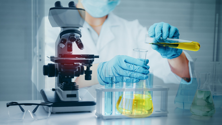 A laboratory technician in a mask, gloves and a microscope pours oil from a test tube into a flask.