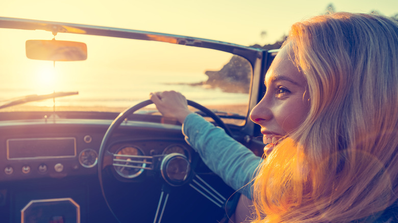 Woman smiling while behind the wheel of a parked classic British roadster at sunset, view looking over left shoulder through windshield