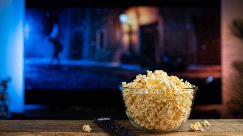 A glass bowl of popcorn and remote control with TV in the background