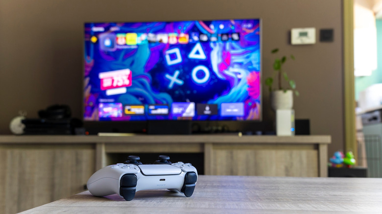 A portrait of a Sony Playstation 5 white basic wireless controller on a wooden table in front of a television showing the PS5 screen.