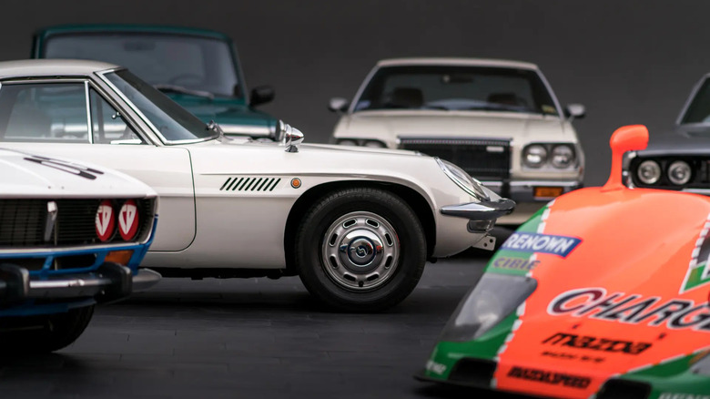 Various Wankel-powered Mazda vehicles including Series I Cosmo Sport, 787B race car, RX-3 race car, Familia R100, and Luce parked together with Cosmo right-side fender in focus