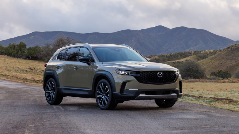 A tan Mazda CX-50 sitting on a road with mountains and hills behind it