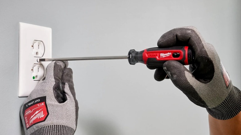 Gloved hands use a 10-inch Milwaukee screwdriver to secure an electrical outlet cover.