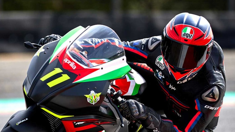 Professional motorcycle racer cornering on a black Aprilia racing bike with Italian livery, wearing AGV helmet and Dainese racing suit during a track day event.