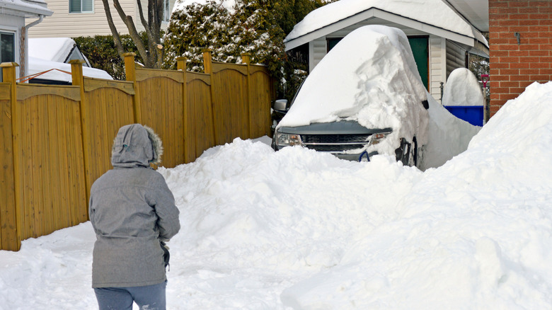 A person in a grey winter coat staring at a driveway, vehicle, and home covered in piles of snow