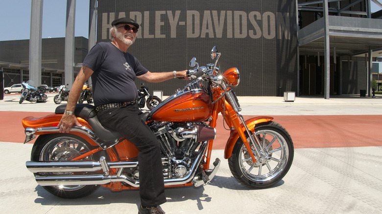 Willie G. poses on an orange motorcycle in front of Harley-Davidson sign