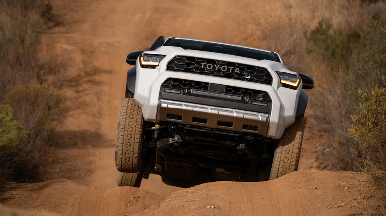 White SUV launching over large bumps on a sandy trail, with scrub on either side. Its undercarriage is visible