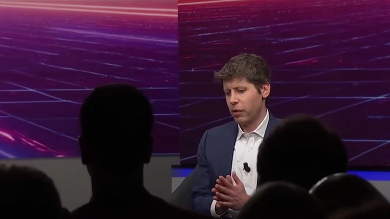 Sam Altman sits in dress shirt and jacket with hands together, abstract neon grid decor behind him