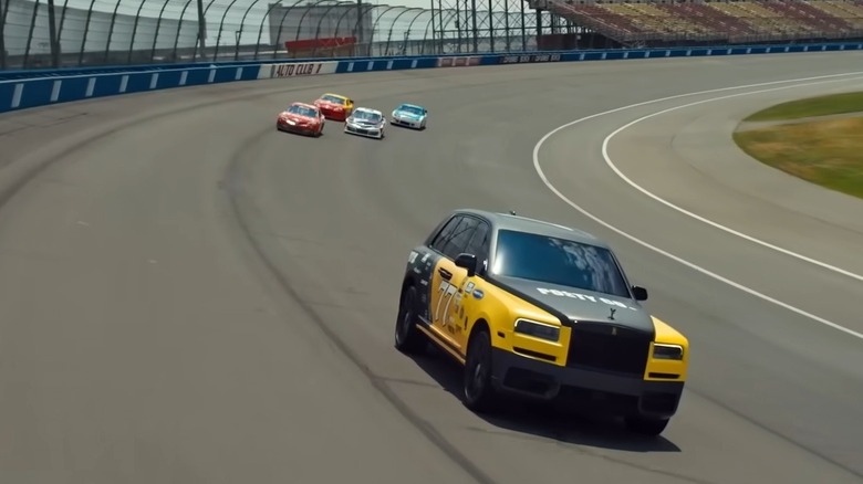 2021 Rolls-Royce Cullinan driving on oval racetrack with stock cars in background, front-right 3/4 view