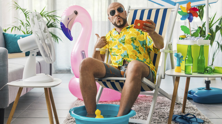 Man in beach wear pretending he's on vacation with feet in a tiny plastic pool, drinks in a cooler, an inflatable flamingo, and more.