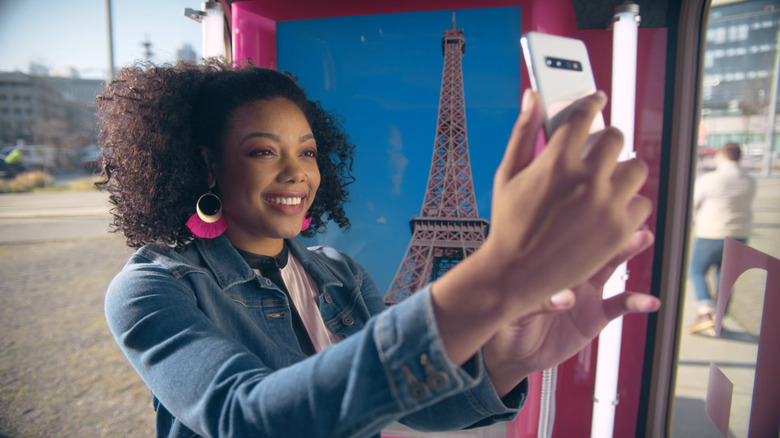 A person holding a white mobile phone up in front of a pink T-Mobile phonebooth and logo branding.