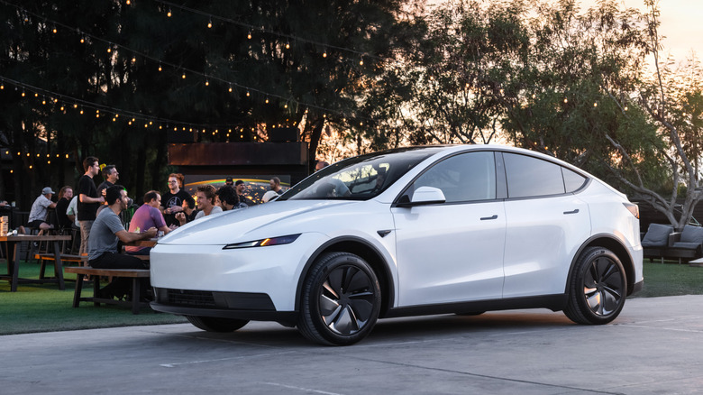 White Tesla Model Y parked outdoors with people sitting at picnic tables in the bakground