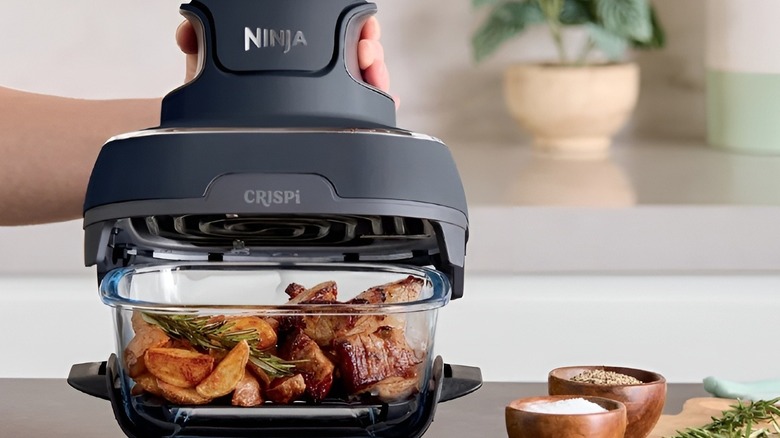 Ninja Crispi air fryer cooking pork and potatoes with rosemary inside a glass container on a kitchen counter