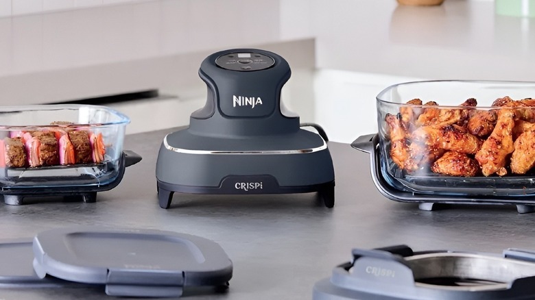 Ninja Crispi air fryer on a countertop between two glass containers, one with chicken wings and one with skewers.