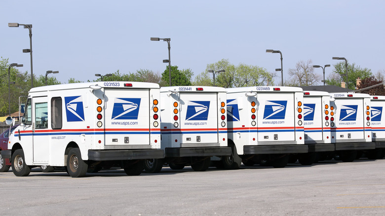 Back view of six United States Postal Service mail delivery vehicles awaiting deployment in Franklin Park, Illinois. They are parked on a lot with blue sky and trees behind them. Their back license numbers are visible.