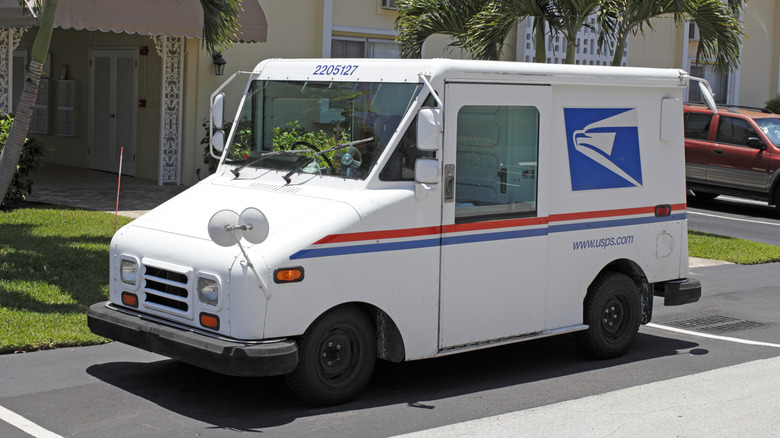 Front side view of a USPS mail truck parked in front of a residential condominium on a sunny day. Its front serial number is clearly visible.