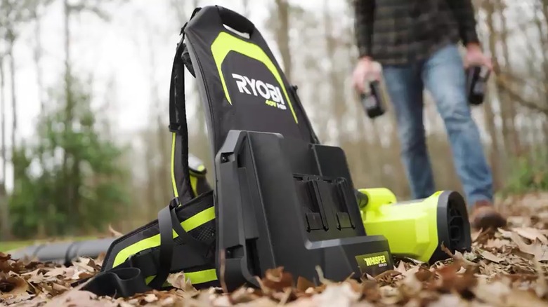 Ryobi backpack blower sits on pile of leaves, man in background approaching with two batteries